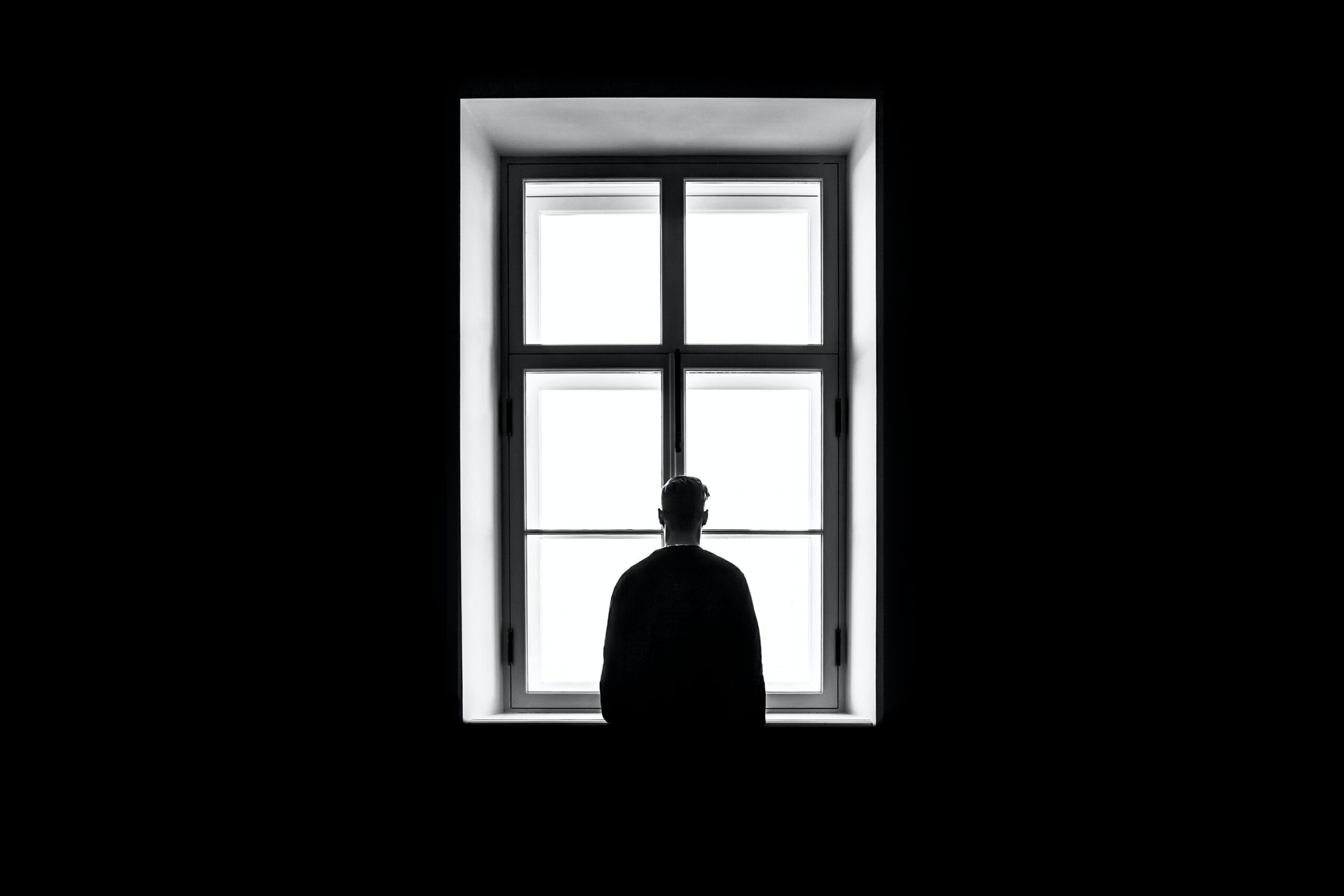 Un homme seul se trouvant dans une pièce très sombre, regarde par la fenêtre